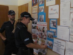 Zdjęcie dwóch funkcjonariuszy Policji umieszczających ulotki na tablicy informacyjnej.