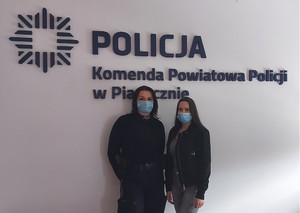 Zdjęcie dwóch kobiet w maseczkach, w tym jedna w umundurowaniu służbowym na tle ściany z logo i Napisem Komenda Powiatowa Policji w Piasecznie.