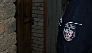 Zdjęcie lewego barku i ręki umundurowanego policjanta na tle ściany piwnicy z czerwonej cegły.