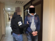 zdjęcie przedstawiające mężczyznę z zasłoniętą twarzą, skutymi z przodu rękoma, przytrzymywanego przez drugiego mężczyznę w korytarzu komisariatu.