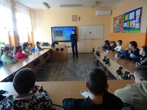 Zdjęcie sali lekcyjnej z uczniami oraz umundurowanym policjantem prowadzącym prelekcję.