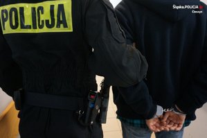 Zdjęcie pleców umundurowanego policjanta i zatrzymanego, skutego z tyłu kajdankami mężczyzny.