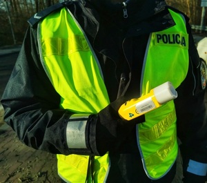 Zdjęcie piersi umundurowanego policjanta w żółtej kamizelce odblaskowej trzymającego w ręce alkotester.