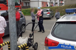 Zdjęcie przedstawiające parking osiedlowy i stojącą na nim policjantkę z dzieckiem na ręku.