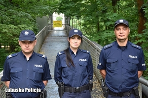 Zdjęcie trójki umundurowanych policjantów, w środku policjantka, na początku mostu w parku.