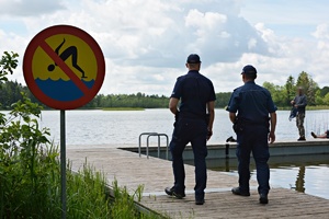 Zdjęcie dwóch umundurowanych policjantów na drewnianym molo jeziora, na pierwszym planie widoczny znak zakaz skoków do wody