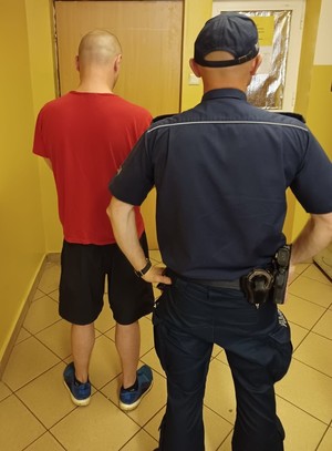 Zdjęcie stojących tyłem umundurowanego policjanta oraz mężczyzny ubranego w krótkie spodenki oraz t-shirt w korytarzu komisariatu.