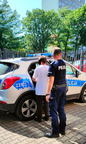 Zdjęcia dziedzińca siedziby Policji, na którym stoi radiowóz, w którym umieszczany jest przez umundurowanego policjanta mężczyzna w białej koszulce.