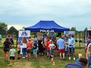 Zdjęcie grupy dzieci i młodzieży na tle zieleni, z tyłu widoczny niebieski namiot wystawienniczy z napisem &quot;Policja&quot;