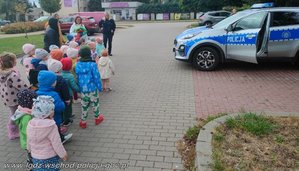 Grupa przedszkolaków na chodniku, na którym stoi policyjny radiowóz.