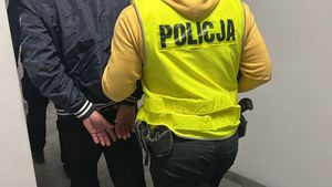 Korytarz komisariatu, widoczne plecy dwóch mężczyzn: ubranego na ciemno z rękami skutymi z tyłu oraz ubranego w żółta kamizelkę Policja.