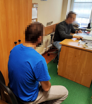 wnętrze pokoju w komisariacie, przy biurku nieumundurowany policjant, na pierwszym planie siedzący tyłem mężczyzna