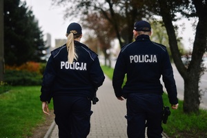 Zdjęcie dwójki umundurowanych policjantów idących chodnikiem wzdłuż pasa zieleni.