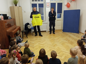 Sala wypełniona siedzącymi na podłodze dziećmi. Przed nimi dwóch umundurowanych policjantów, z których jeden w ręku trzyma żółtą kamizelkę odblaskową.