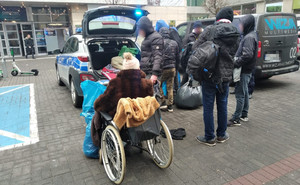 Parking uliczny, zaparkowany radiowóz z otwartym bagażnikiem, otoczony grupą ludzi, w tym kobieta na wózku inwalidzkim.