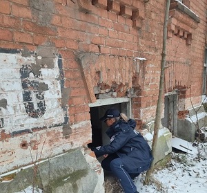 Ściana starego budynku z czerwonej cegły, z otworem w dolnej części, przy którym kuca umundurowana policjantka