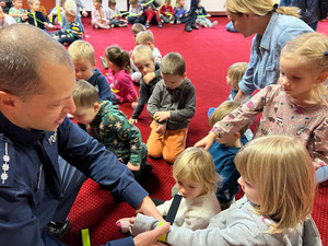 Sala przedszkolna z siedzącą na podłodze grupką dzieci, wśród nich umundurowany policjant.