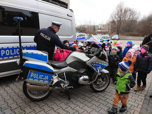 Parking Policji, w tle radiowozy, na pierwszym planie motocykl i umundurowany policjant otoczony grupką dzieci.