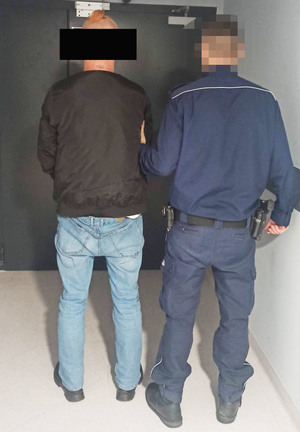 Stojący tyłem przy ścianie mężczyzna przytrzymywany przez umundurowanego policjanta.
