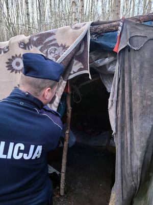 Umundurowany policjant zaglądający do prowizorycznego szałasu z kocy na tle lasu
