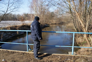 Umundurowany policjant stojący na mostku nad rzeką w scenerii zimowej.