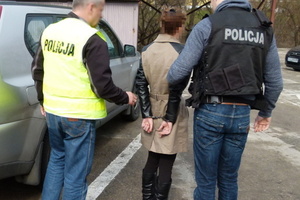 Kobieta ze skutymi z tyłu rękami przytrzymywana przez dwóch nieumundurowanych policjantów ubranych w kamizelki z napisem policja.