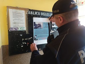 Umundurowany policjant przypinający kartkę na tablicy ogłoszeń wewnątrz budynku.