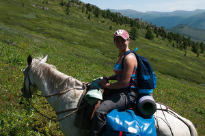 Kobieta jadąca na białym koniu stokiem górskim porośniętym zieloną trawą.