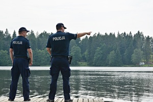 Dwaj umundurowani, odwróceni tyłem, stojący na molo nad jeziorem policjanci