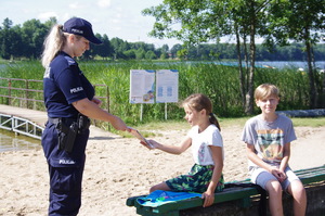 Dwójka dzieci siedząca na ławce, policjantka wręczająca ulotkę, w tle jezioro