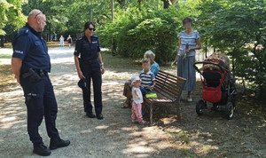 Seniorka siedząca na ławce w parku z dwójką dzieci, obok dwójka umundurowanych policjantów.