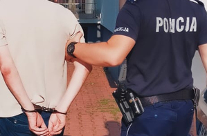 Zbliżenie pleców mężczyzny z zakutymi z tyłu rękami przytrzymywanego przez umundurowanego policjanta.