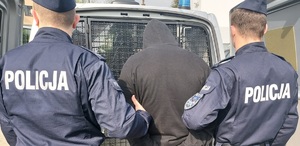 Zbliżenie widzianych od tyłu: mężczyzny ubranego w czarną bluzę z kapturem ze skutymi z tyłu rękami przytrzymywanego przez dwóch mundurowych policjantów na tle krat radiowozu