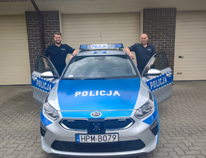Dwóch uśmiechniętych umundurowanych policjantów stojących po obu bokach radiowozu