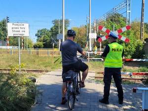 Przejście kolejowe przy którym odwróceni tyłem stoją: rowerzysta oraz policjant