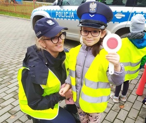 Dziewczynka w czapce policyjnej i kamizelce odblaskowej obok policjantki