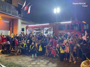 W scenerii nocnej grupa dzieci na tle wozu strażackiego, widoczne osoby dorosłe w mundurach rożnych formacji