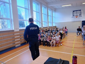 Sala gimnastyczna wypełniona dziećmi oraz policjant prowadzący pogadankę