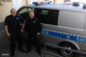 Zdjęcie przedstawiające dwójkę policjantów stojących przy oznakowanym radiowozie policyjnym