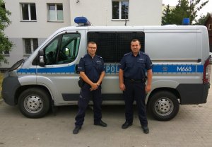 Zdjęcie przedstawiające dwójkę umundurowanych policjantów stojących koło oznakowanego radiowozu policyjnego