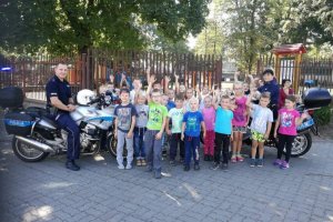 Zdjęcie przedstawiające policjanta siedzącego na motocyklu służbowym i stojącymi obok niego przedszkolakami