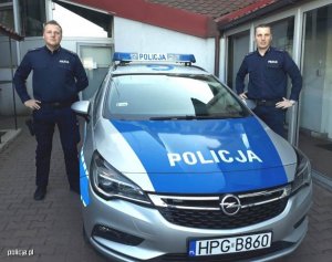 Zdjęcie przedstawiające dwójkę policjantów  stojących obok radiowozu