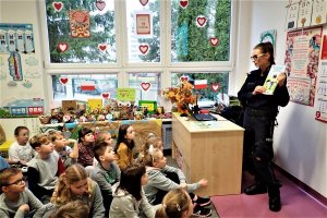 Zdjęcie przedstawiające policjantkę stojącą naprzeciw dzieci w klasie, trzymającą w ręku książkę edukacyjną.