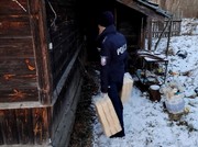 Zdjęcie umundurowanego policjanta odwróconego tyłem na tle drewnianego domu w scenerii zimowej.
