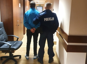 Zdjęcie w korytarzu komisariatu przedstawiające mężczyznę odwróconego tyłem z kajdankami założonymi na ręce trzymane za tułowiem oraz również odwróconego tyłem umundurowanego policjanta.