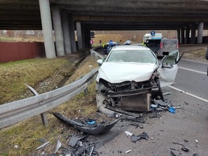 Zdjęcie samochodu osobowego z uszkodzoną przednią nadwozia na tle dolnej części wiaduktu drogowego.