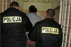 Zdjęcie przedstawiające mężczyznę odwróconego plecami w pomieszczeniu aresztu przytrzymywanego przez dwóch umundurowanych policjantów.