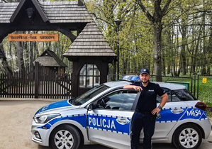 Zdjęcie przedstawiające umundurowanego policjanta stojącego  w otwartych drzwi oznakowanego radiowozu na tle drewnianej bramy wjazdowej Białobrzeskiego Parku Narodowego.