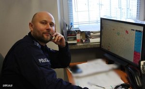 zdjęcie umundurowanego policjanta siedzącego przy biurku służbowym w tle widoczny monitor komputerowy na którym wyświetlona jest KMZB.