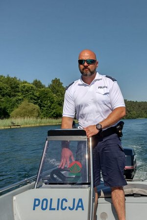 Zdjęcie umundurowanego w strój wodniaka policjanta (krótkie granatowe spodnie, biała koszulka) na łodzi motorowej.